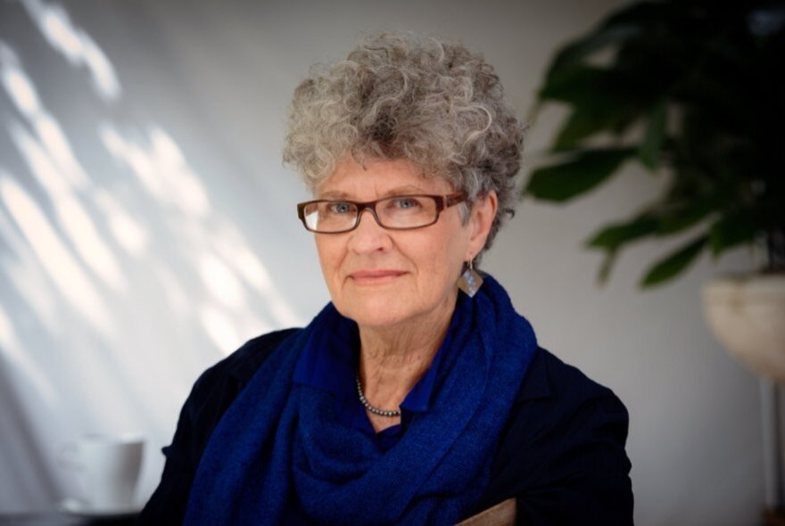 Author Kate Grenville, with grey hair and spectacles, in a shady room with plants.
