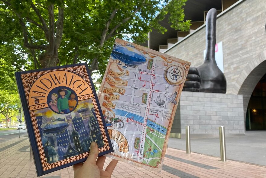 A hand holding up a map and a book in a garden setting with a large sculpture visible. 