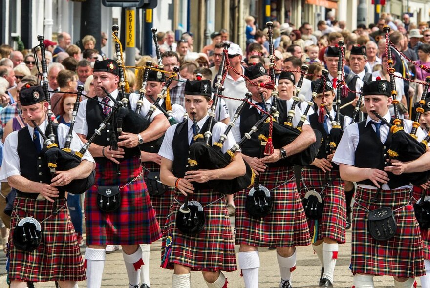 A parade of men wearing kilts, walking in formation down Collins Street, playing the bagpipes, with onlookers in the background.