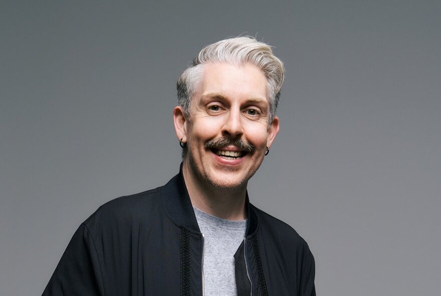 Portrait of Australian comedian, Carl Donnelly, smiling and wearing a black bomber jacket; grey background.