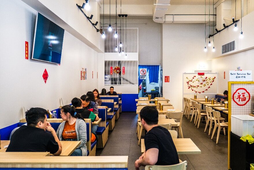 The crisp, white interior of an Asian cafe, with people seated at wooden tables with booth seating and vibrant blue paint on the lower portion of the walls.