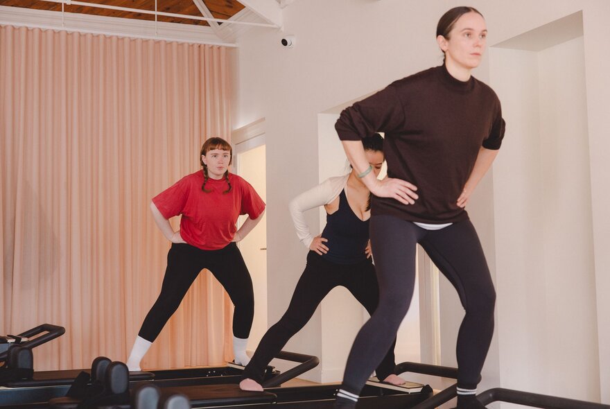 A reformer Pilates class with people on machines with hands on hips.