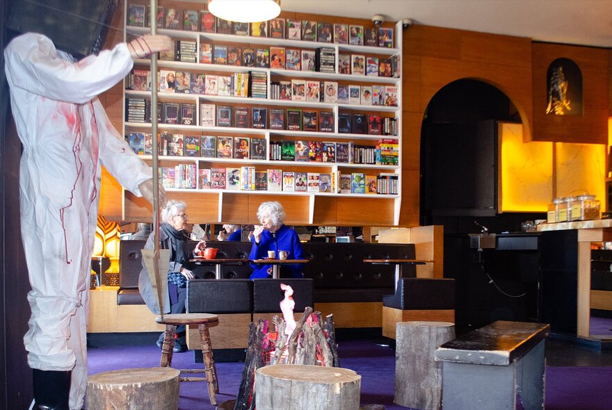 Person wearing blood-spattered white dungarees in a bar with shelves and props.