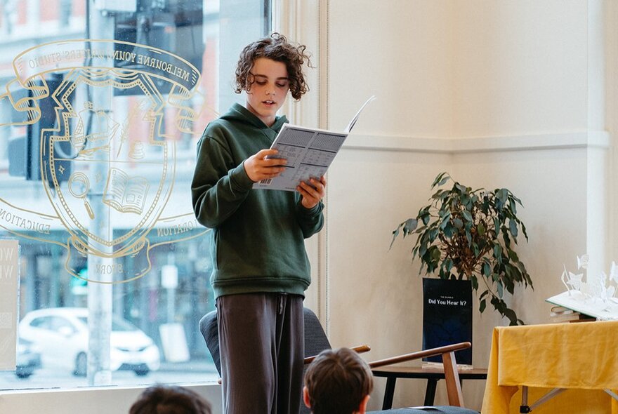 A kid standing up in front a class reading in front of a large window. 