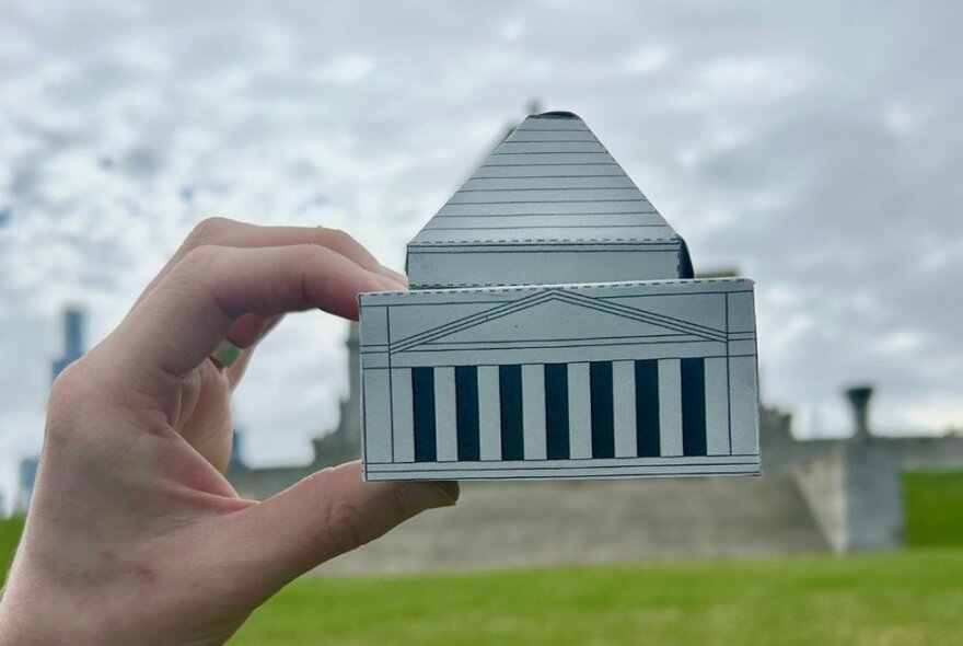 A hand holding a cardboard model of the Shrine of Remembrance.