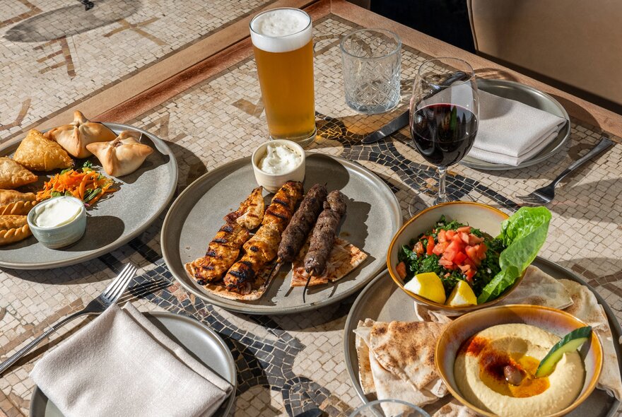 Overhead view of a table in a restaurant with a plate of dips, bread and salad, meat skewers, napkins, cutlery, a glass of wine and a beer.