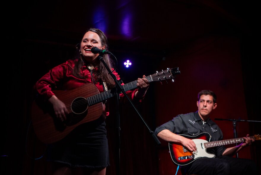 A woman and man both playing guitar on a stage. 