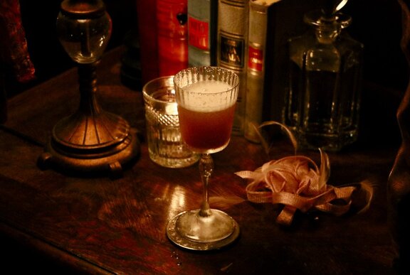 A bellini in an ornate glass on a table with books and a candle. 