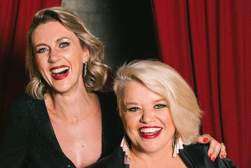 Singers Amelia Ryan and Libby O’Donovan, both wearing dark tops and smiling, posed against a red velvet curtain.