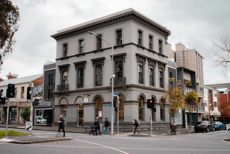 The exterior of a three level 19th century Victorian style building on a street corner, with an intersection, pedestrians and parked cars visible.