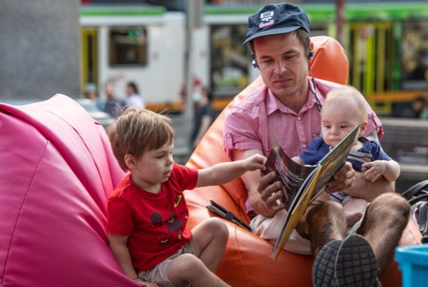 Adult male with two children reading on beanbags with a tram in the background.