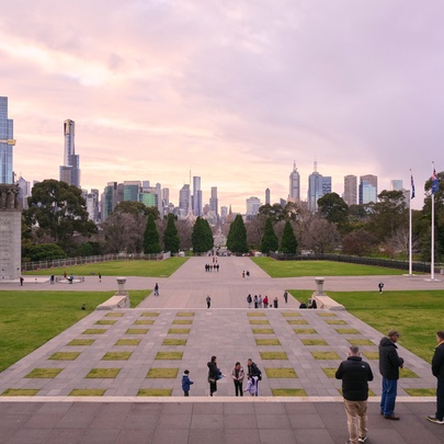 A wide-angle photo taken from the top steps of the Shrine of Remembrance in Melbourne, looking down a paved forecourt and across parkland towards the city skyline at dusk. 