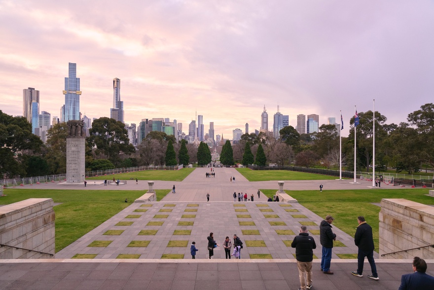 A wide-angle photo taken from the top steps of the Shrine of Remembrance in Melbourne, looking down a paved forecourt and across parkland towards the city skyline at dusk. 