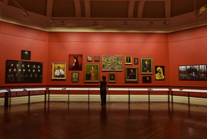 Red walled gallery space in the State Library of Victoria with a single person looking at paintings on the wall. 