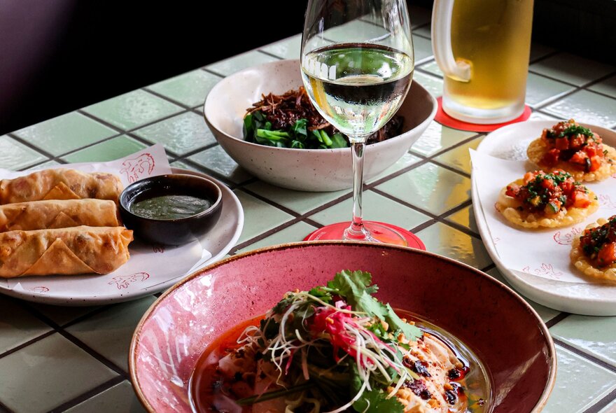 Several dishes of Asian food on a tiled table with a glass of white wine.