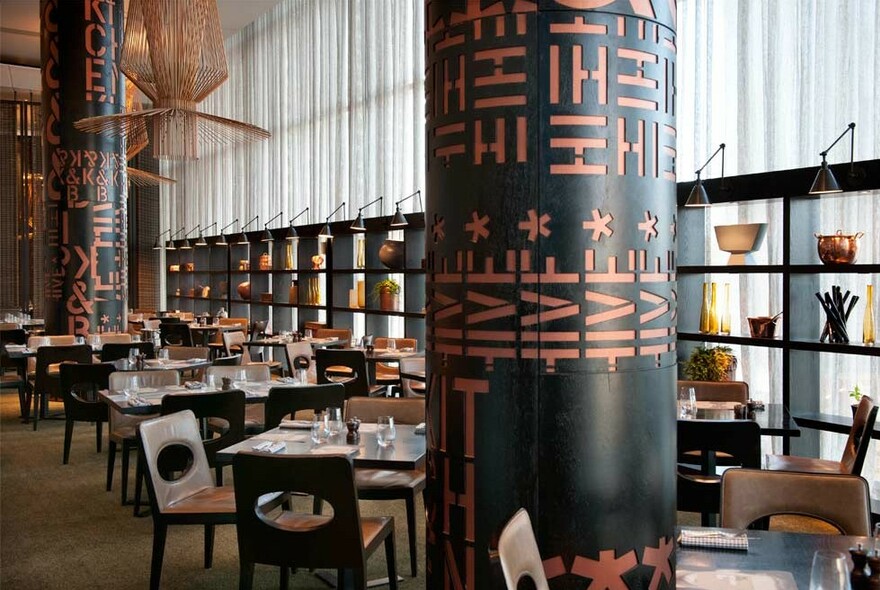 Formal dining room with tables and chairs, light curtained windows, backlit shelving and large black and copper pillars.