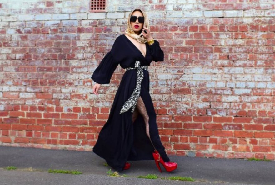 Model wearing a long black open coat with tie, sunglasses, scarf and red platform shoes.