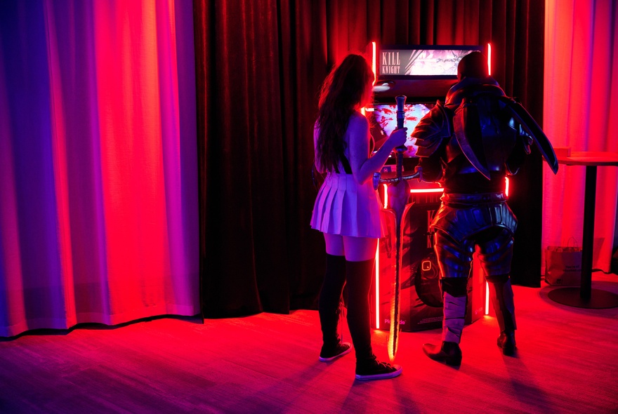 Two figures wearing gaming costumes standing at an illuminated games machine.