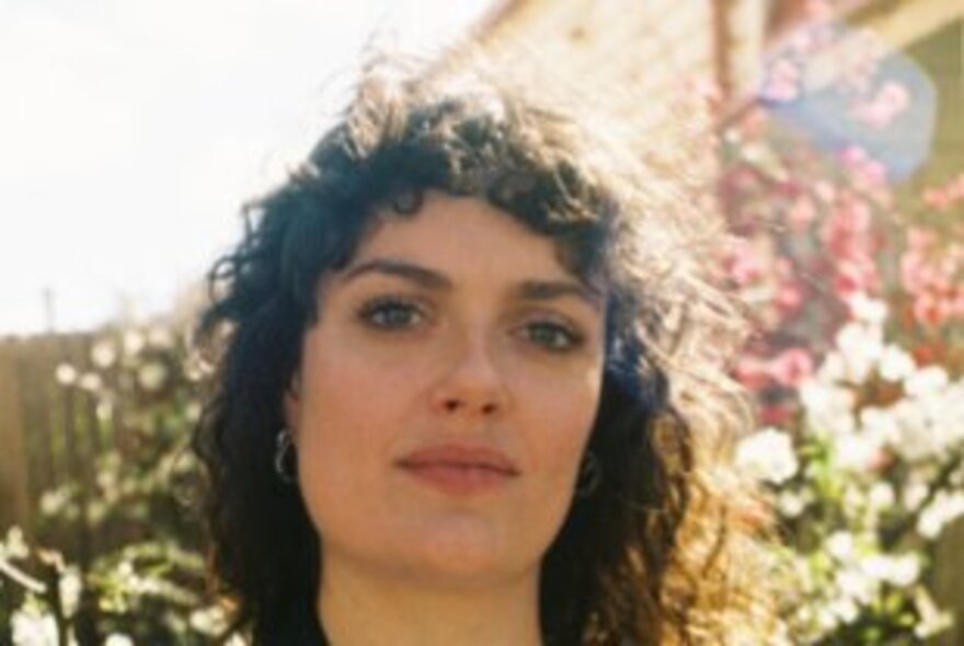 A close-up portrait of a person with curly brown hair, smiling faintly while standing outdoors with a bright, sunlit floral background.