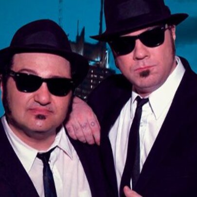 Two performers portraying Jake and Elwood Blues, famously known as The Blues Brothers, wearing dark sunglasses, black suits and ties, white shirts and black pork pie hats, posed against a city skyscraper background.