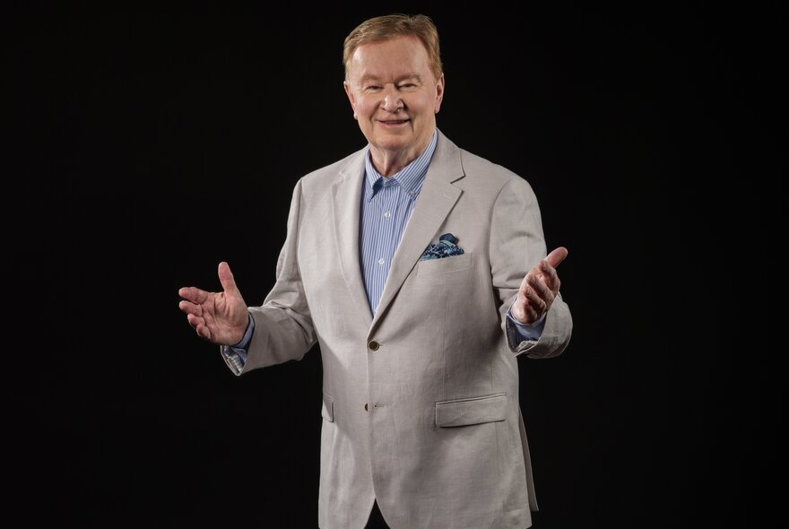Australian personality, Denis Walter, wearing a grey suit with arms outstretched in a welcoming gesture; black background.