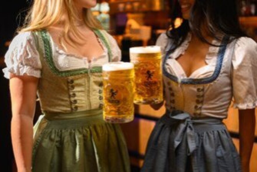 Two women wearing traditional Bavarian dirndls and holding large glass mugs of beer in a bar.