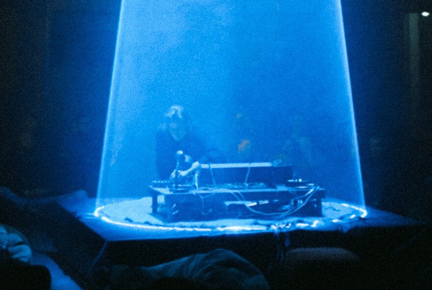 A blue cone spotlight used during a live performance, lighting up a performer at an electronic keyboard or similar instrument, with an audience listening in the dark.