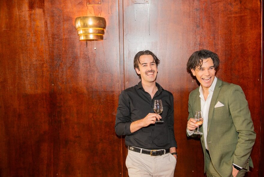 Two men holding glasses of wine and smiling in a room with wood panelled walls.