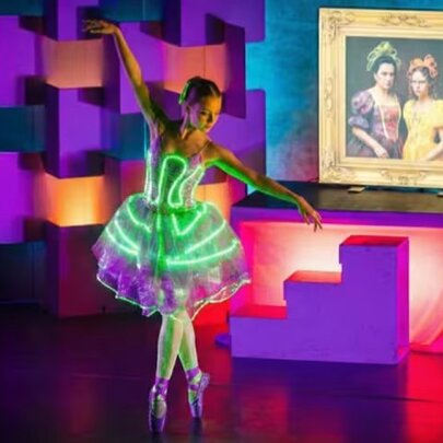 A dancer performs on a brightly illuminated stage wearing a white tutu and bodice outlined with glowing green neon lights, behind her is a framed portrait of two women.
