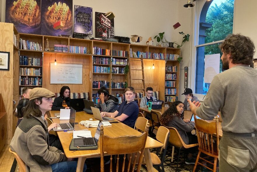 Teenagers sitting around large work tables,  interacting with each other and their teacher in a large room, with windows looking out onto the street and a wall of bookshelves in the back of the room.