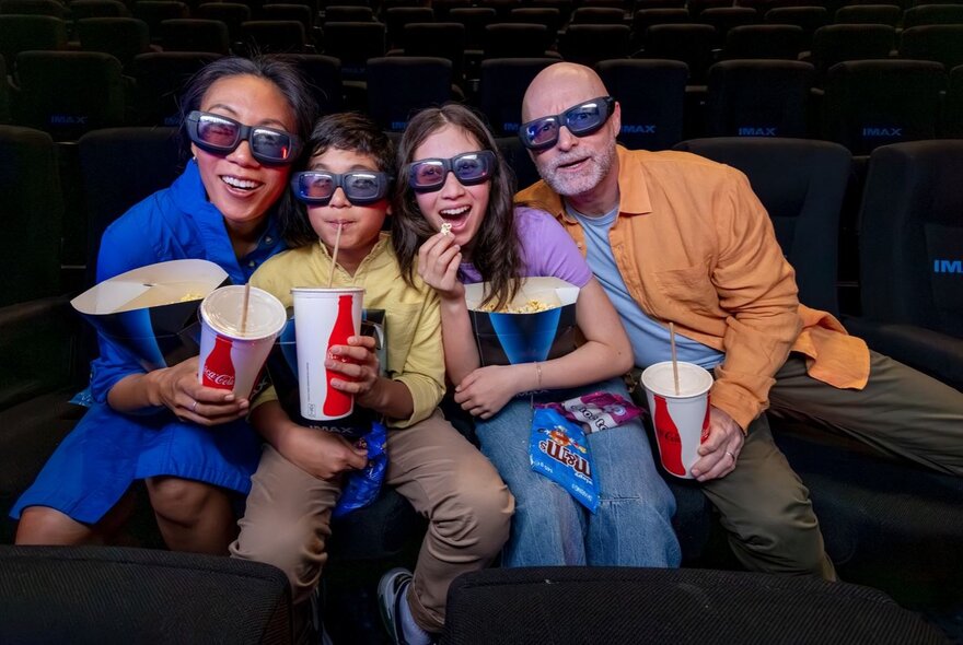 A family of four wearing sunglasses, holding boxes of popcorn and soft drinks.