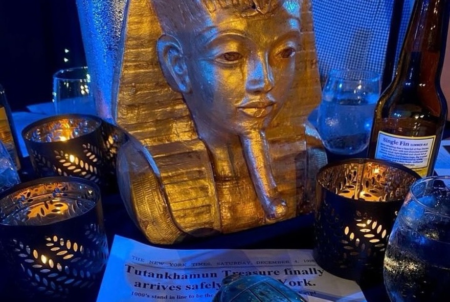 Props on display on a table at a murder mystery dinner show that include a gold bust of Tutankhamun, a flyer, and glasses of wine.