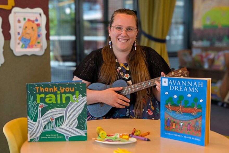 A smiling woman with a ukulele seated at a desk with two picture books on display. 