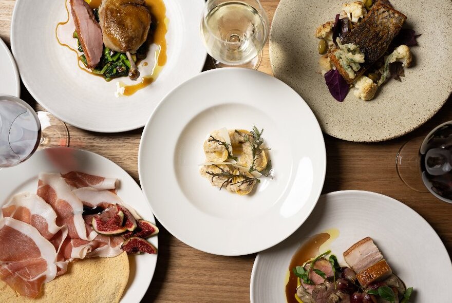 White plates with a variety of dishes on wooden table, glasses of wine visible - from above.