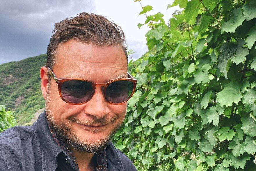 A man in sunglasses in front of a lush grape vine, with hills and clouds in the distance.