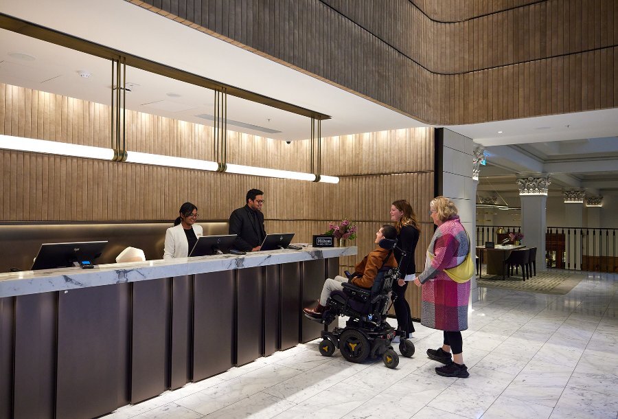 Three people checking in at a hotel lobby, one person is using a wheelchair.