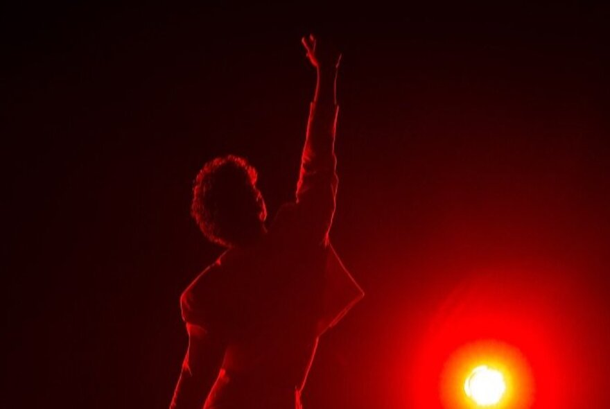 A performer with a single arm raised and facing away from the camera, backlit by a bright red light.