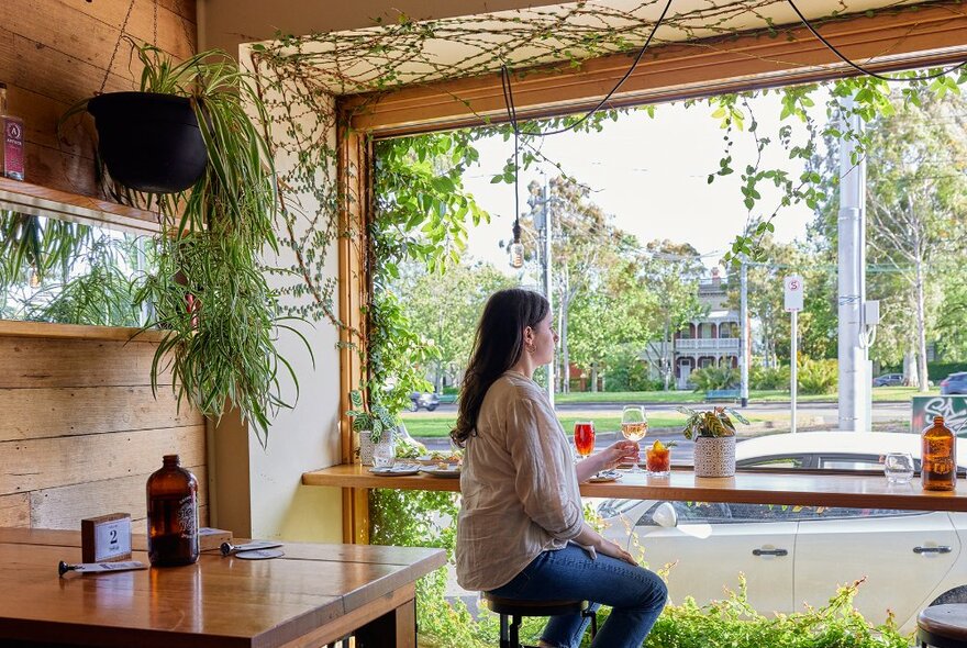 A woman sitting at a window bar seat in a leafy bar.