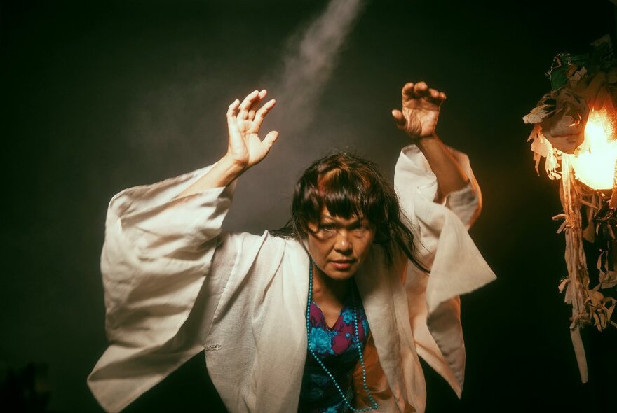 A woman wearing a colourful dress under a white cape with her arms in the air, a fiery talisman hanging beside her.