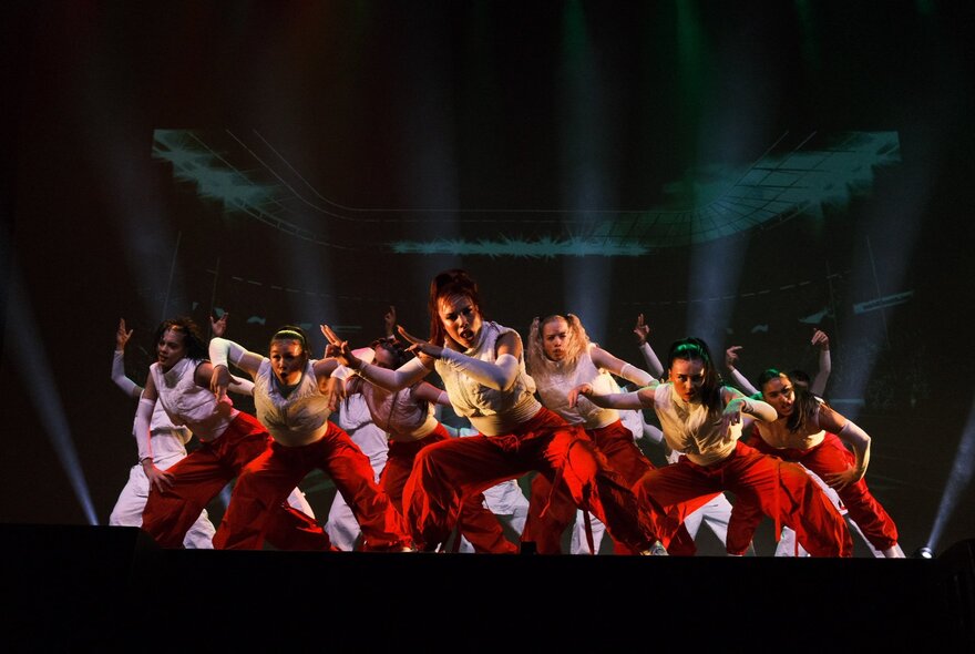 A large group of dancers from the Royal Family Dance Crew, in white tops and red pants perform a high-energy routine on a dark stage with dramatic lighting.