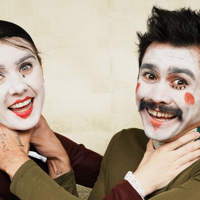 A woman and man wearing clown makeup, smiling, with their hands around each other's throats. 