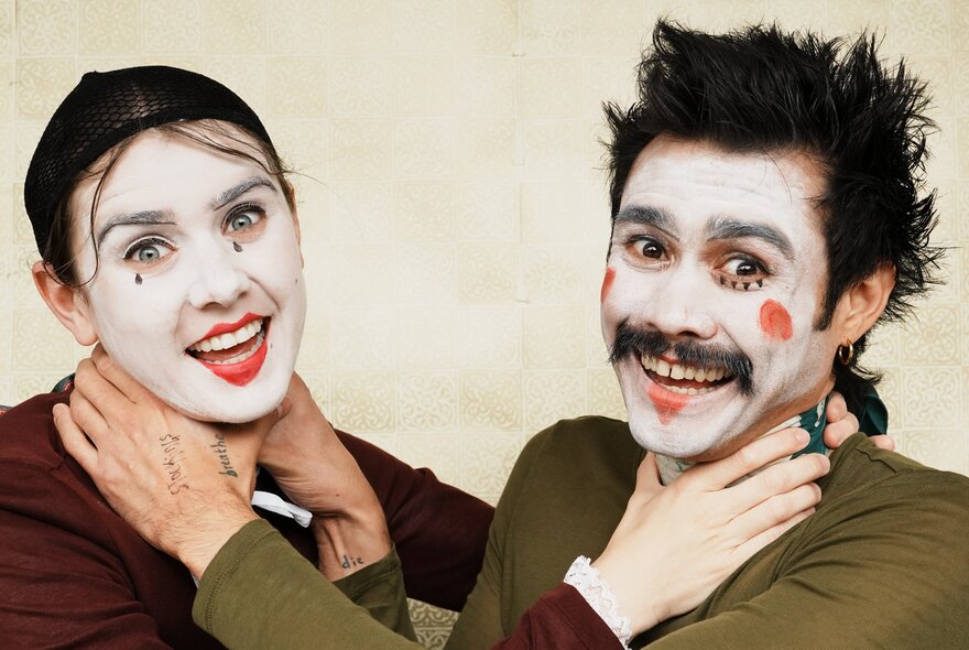 A woman and man wearing clown makeup, smiling, with their hands around each other's throats. 