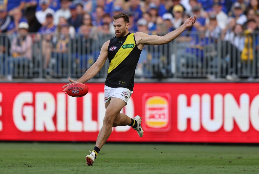 A Richmond AFL football player just about to kick the footy on the field during a match, a blurred crowd of stadium fans in the background.