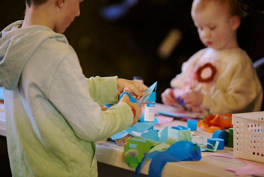 Kids busy at a crafting table, the older one using scissors. 