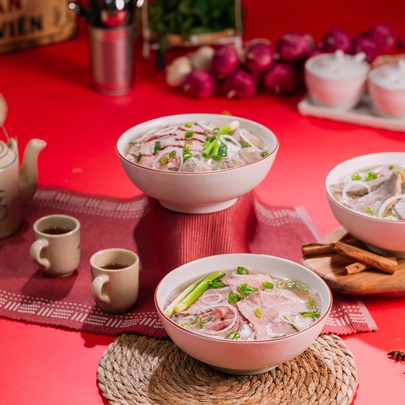 Three bowls of pho, a teapot and cups on a red hued table setting. 