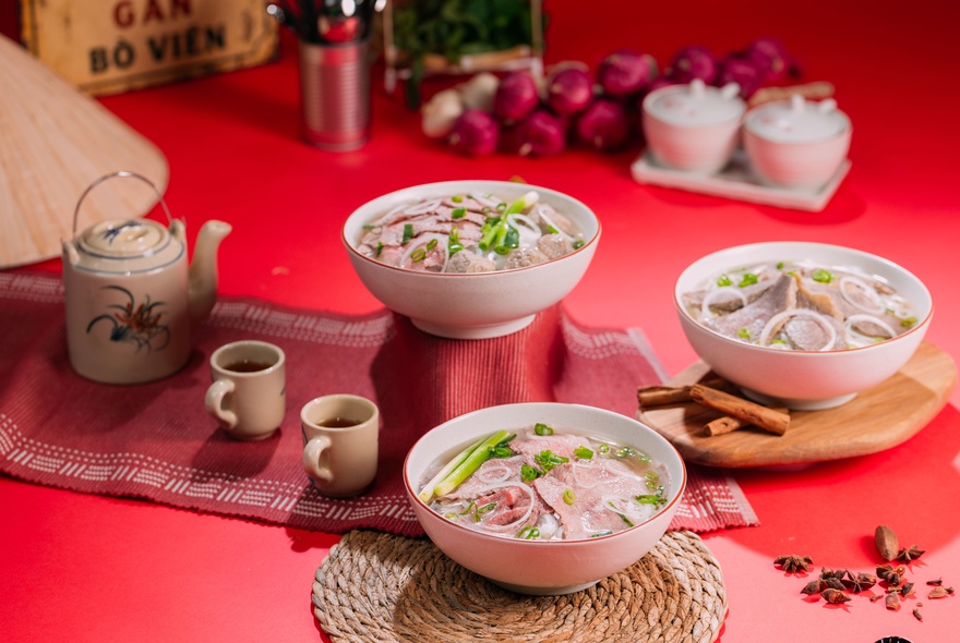 Three bowls of pho, a teapot and cups on a red hued table setting. 