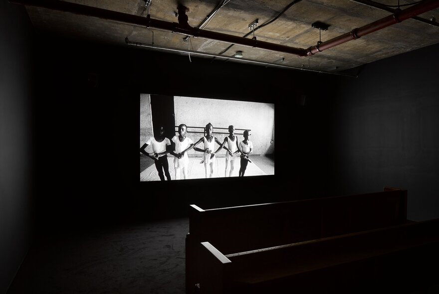 Installation at The Vinyl Factory: Reverb exhibition of a black-and-white video projection on a large screen in a dark exhibition room with benches visible in the foreground for viewers.