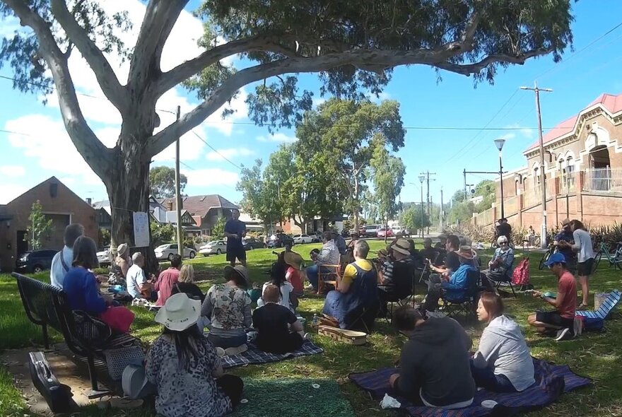 Ukulele group seated with instruments under a tree in a community garden setting.