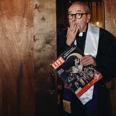 A man wearing clerical vestments holding a vintage LIFE magazine and covering his mouth with his hand as if shocked or surprised.