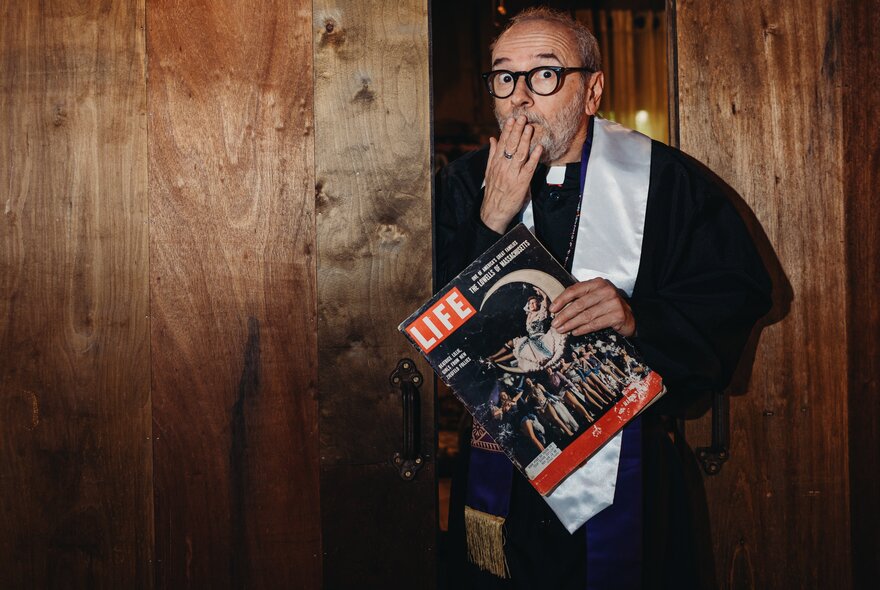 A man wearing clerical vestments holding a vintage LIFE magazine and covering his mouth with his hand as if shocked or surprised.